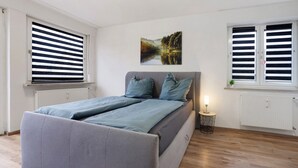 A bedroom with a bed, two windows with blinds, a bedside table with a lamp, and a wall-mounted picture. - Apartment 'Allgäu Space' with Shared Garden, Balcony and Wi-Fi (Marktoberdorf)