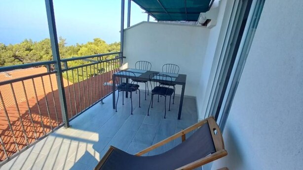 A balcony with a table and chairs, a wooden chair, and a view of greenery. - Berti Sun & Sea Apartments (Hvar)