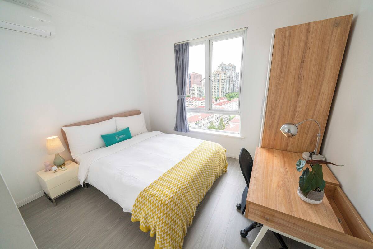 A hotel room with a bed, bedside table, lamp, and a view of buildings through the window.