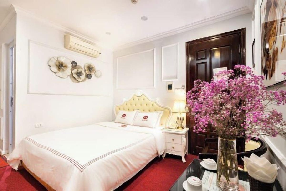 A hotel room with a bed, bedside table, lamp, and a vase with flowers.
