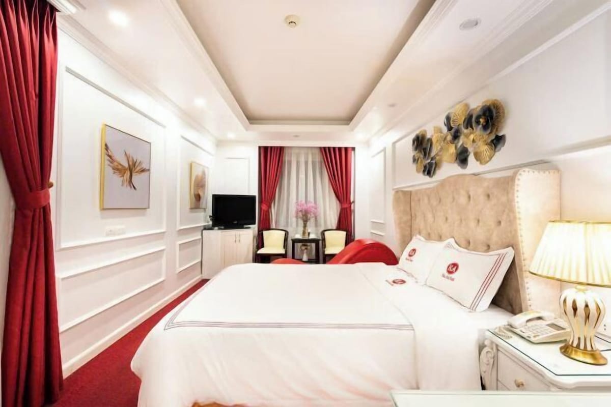 A hotel room with a large bed, a bedside table with a lamp, a TV, and a seating area with chairs and a table.