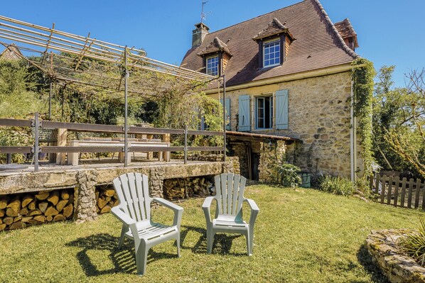 Une maison en pierre avec un toit de tuiles, un mur en pierre avec un foyer, et deux chaises en plastique blanches au premier plan.