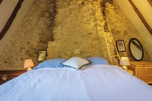 Une chambre avec un mur en pierre, un lit avec une literie bleue, deux lampes de chevet, une échelle en bois, une commode en osier et un tableau encadré accroché au mur.