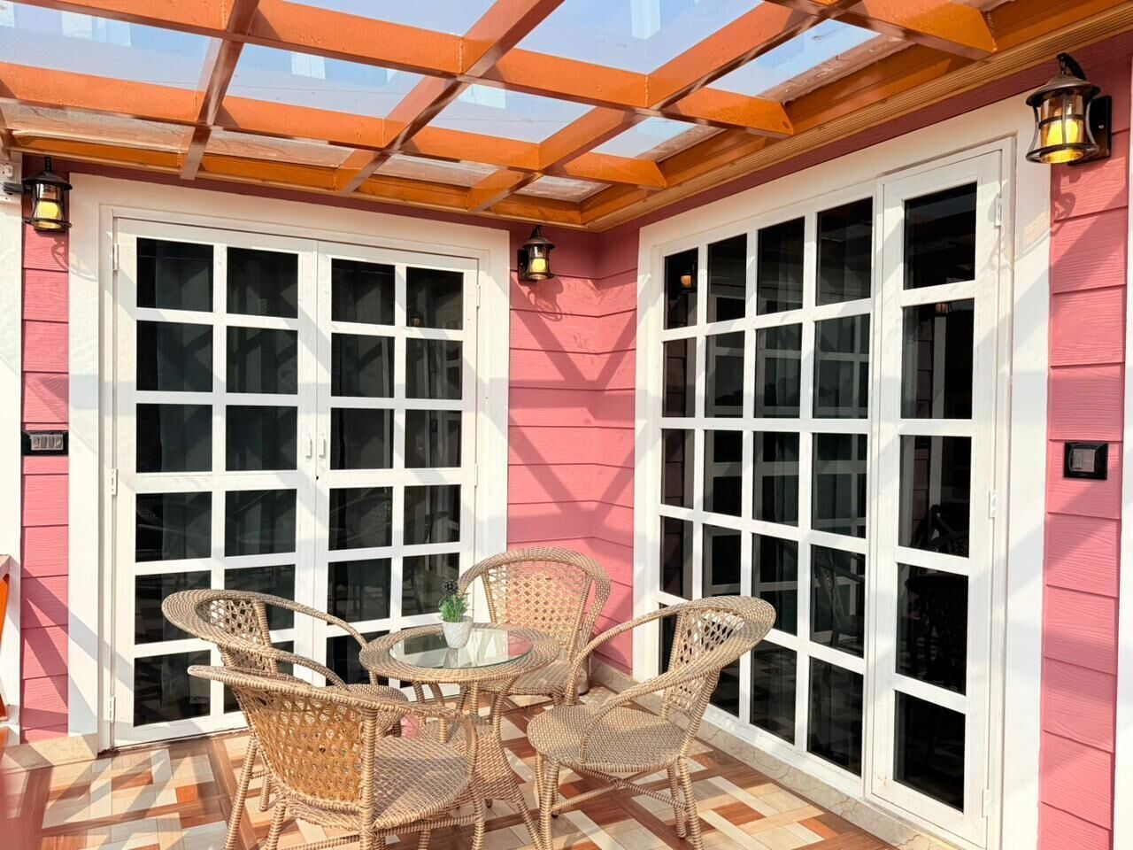 A covered patio with wicker furniture and a glass table, adjacent to a building with pink siding and white-framed windows.