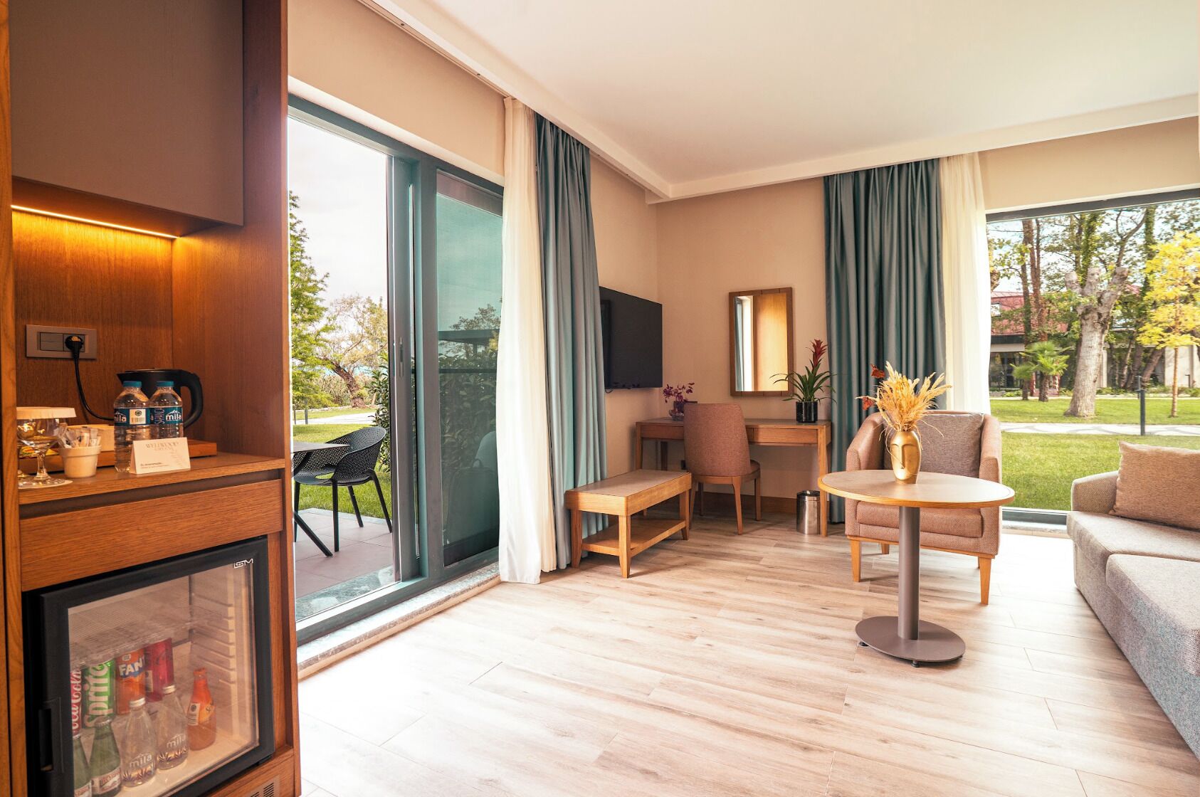 A modern hotel room with a large sliding glass door to the outdoors, a wooden desk, a sofa, and a small round table.