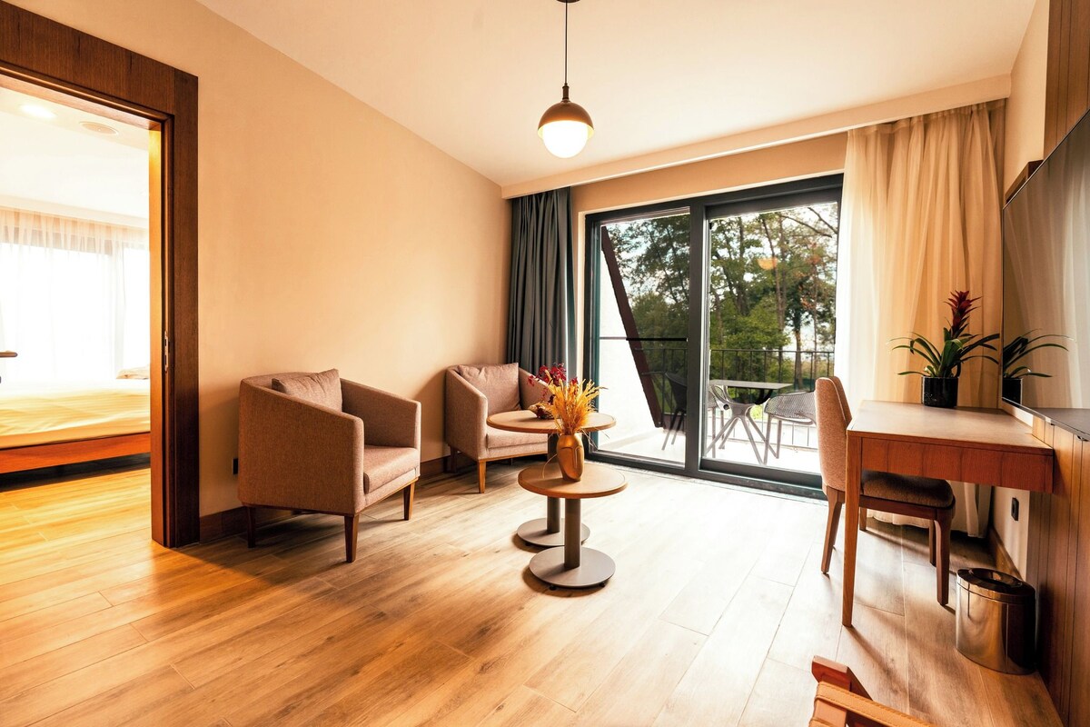 Une chambre moderne avec un plancher en bois, un bureau, deux chaises, une petite table avec un vase et une grande fenêtre avec des rideaux.