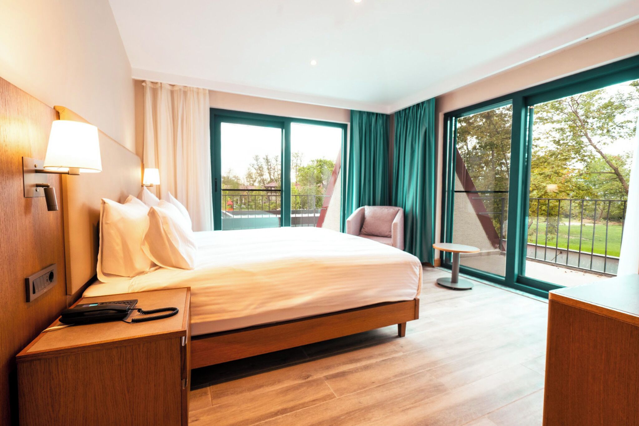 A hotel room with a bed, bedside table, lamp, and a view of the outdoors through large windows.