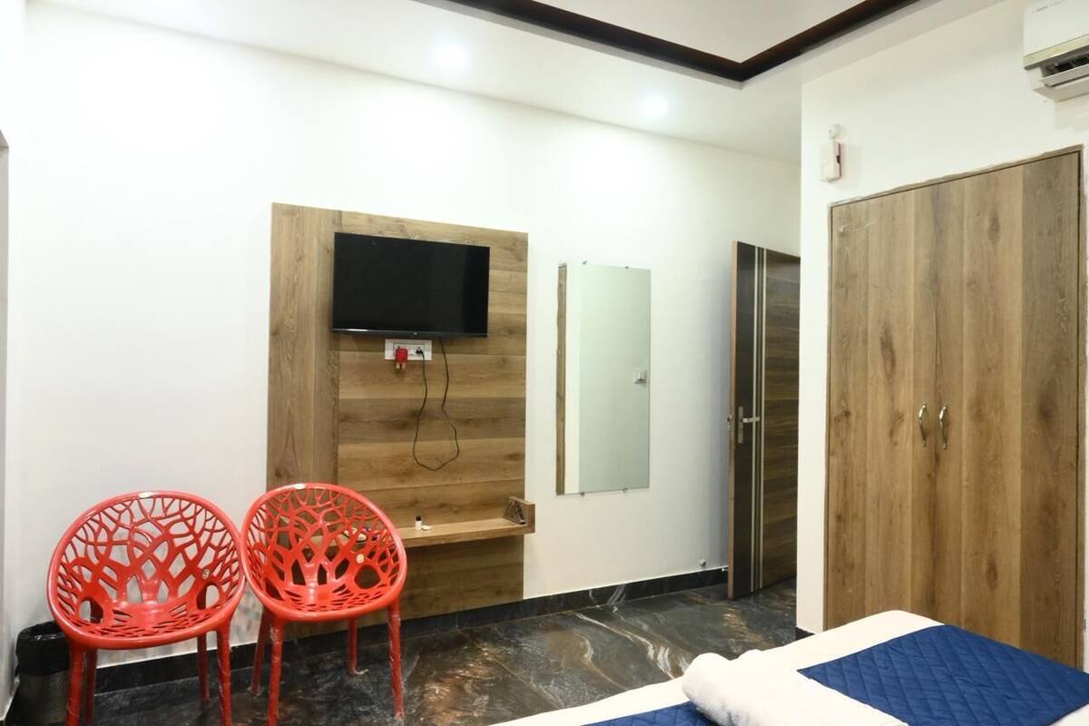 A hotel room with a wooden wall, a flat-screen TV, a mirror, a wardrobe, and two red chairs.