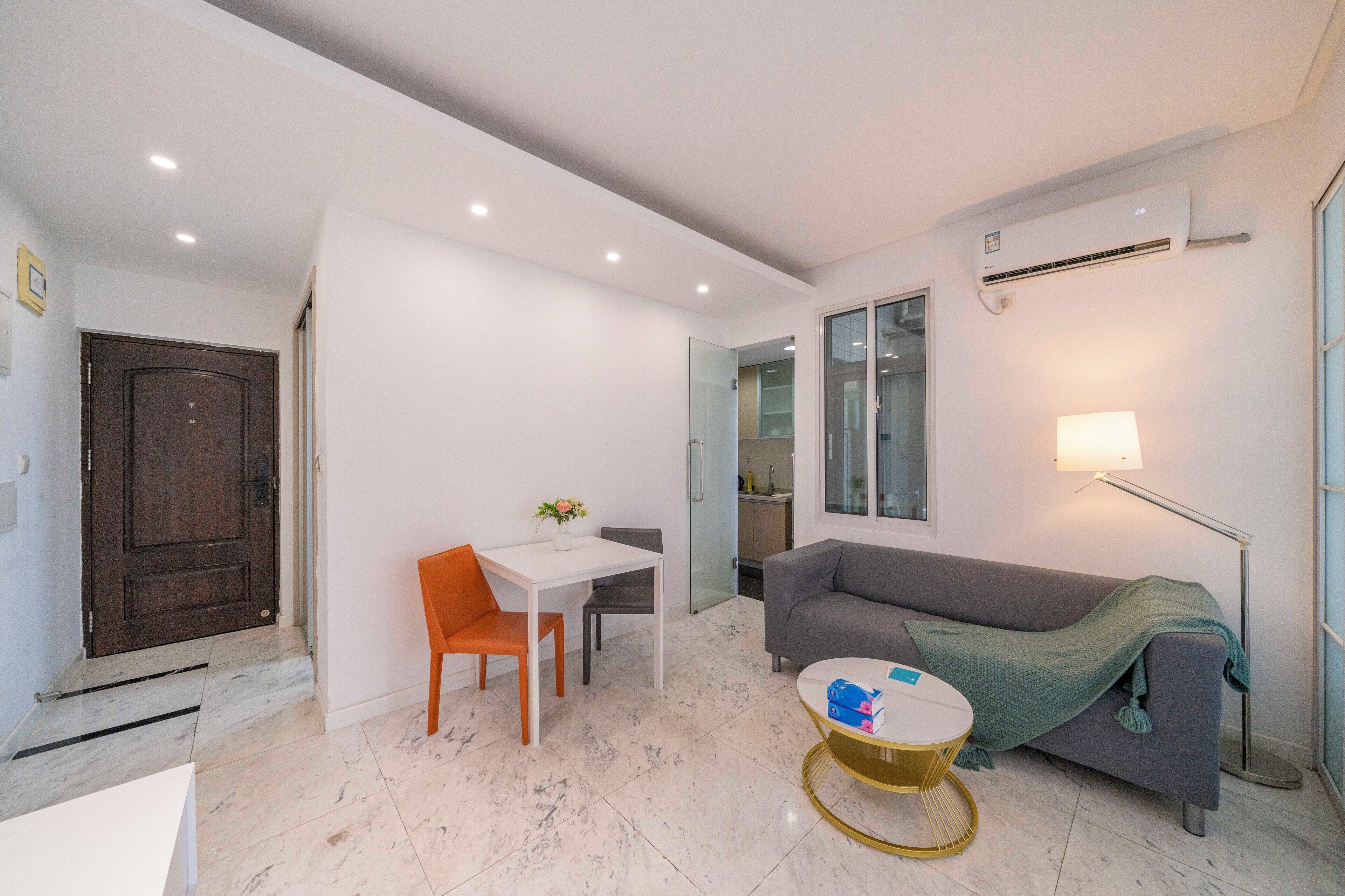 Una sala moderna con una mesa blanca, sillas anaranjadas, un sofá gris y una lámpara de pie.