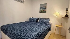 Ein Schlafzimmer mit einem Bett, einer Wandklimaanlage, einem Gemälde, einer Nachttischlampe und einem kleinen Tisch mit Lampe.