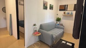 Ein kleines, modernes Wohnzimmer mit einem grauen Sofa, einem kleinen Beistelltisch mit Buch und Pflanze sowie einer Wandregal mit Dekoration.