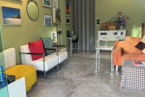 Een zithoek met een witte bank, een geel poefje en een geruit poefje. Er staat een witte salontafel en een witte kast met decoratieve accessoires.