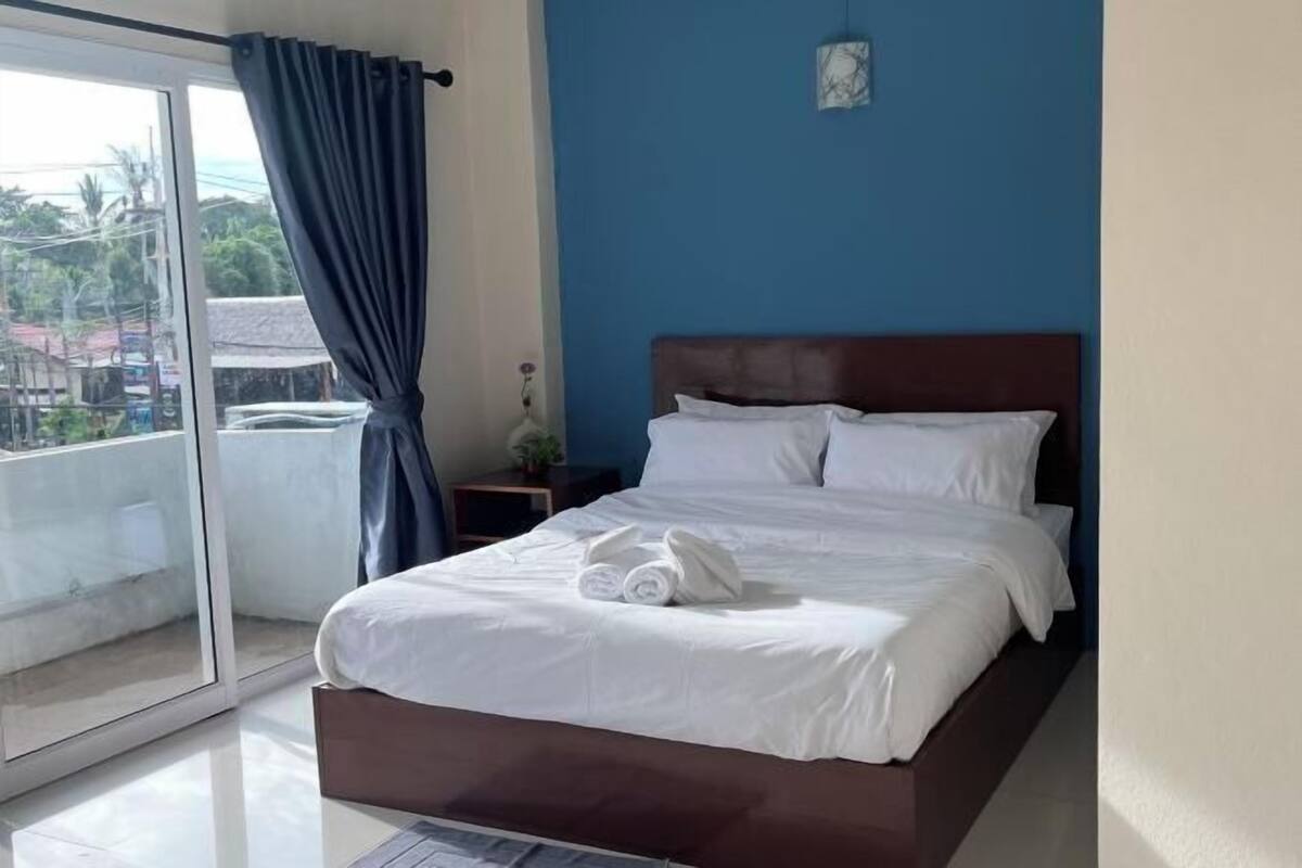Un lit bien fait, avec du linge de lit et des serviettes blanches, une tête de lit marron foncé et une vue sur l’extérieur par une porte vitrée.