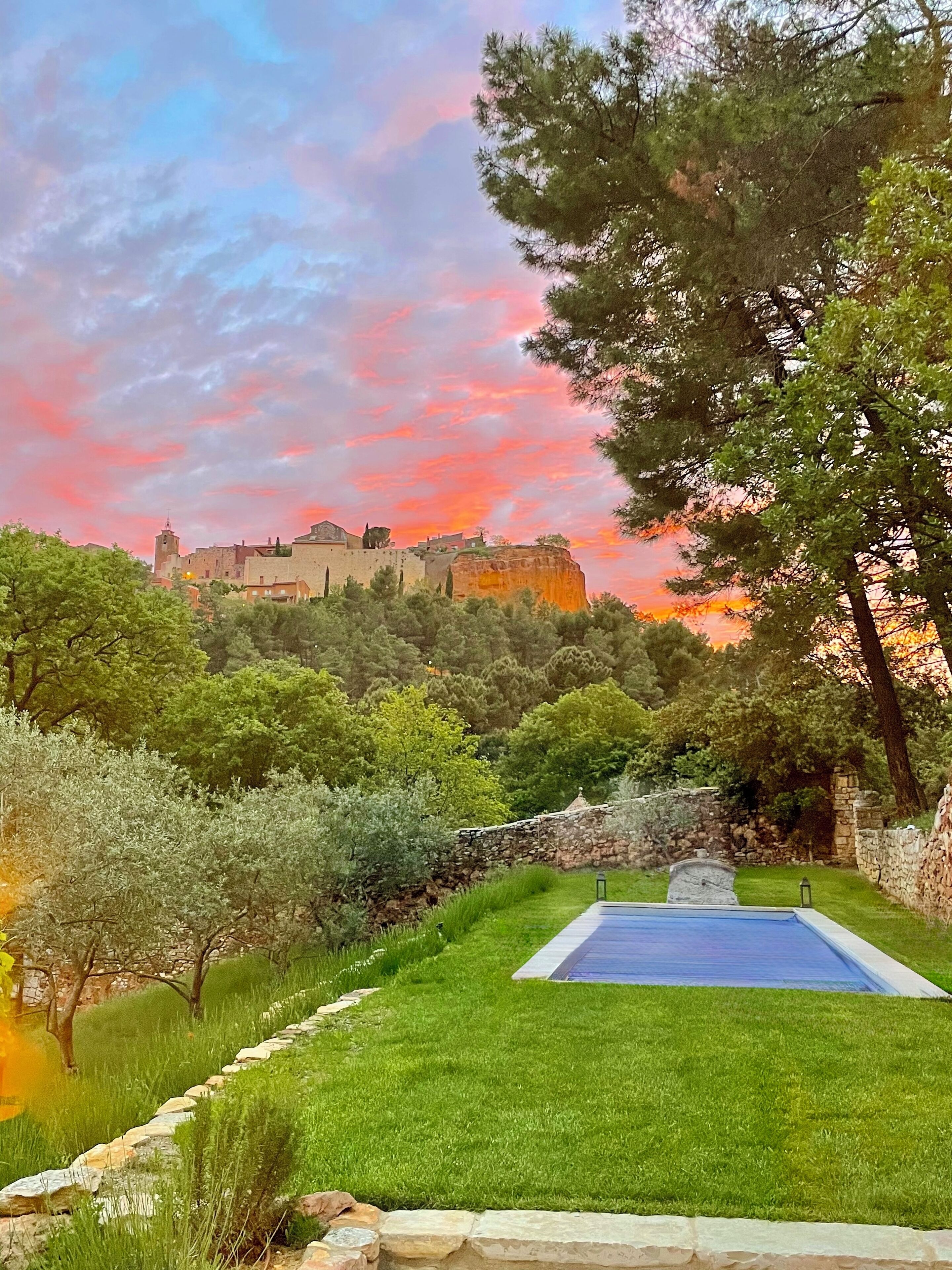 A swimming pool surrounded by a stone wall, lush greenery, and a historic building in the background.