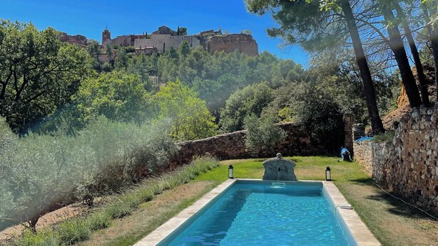 Una piscina rectangular con borde de piedra, rodeada de césped cuidado y árboles, con vista a un edificio histórico al fondo.