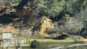 Un camino de grava que lleva a una pequeña construcción con puerta azul, rodeada de árboles y un terreno montañoso rocoso.