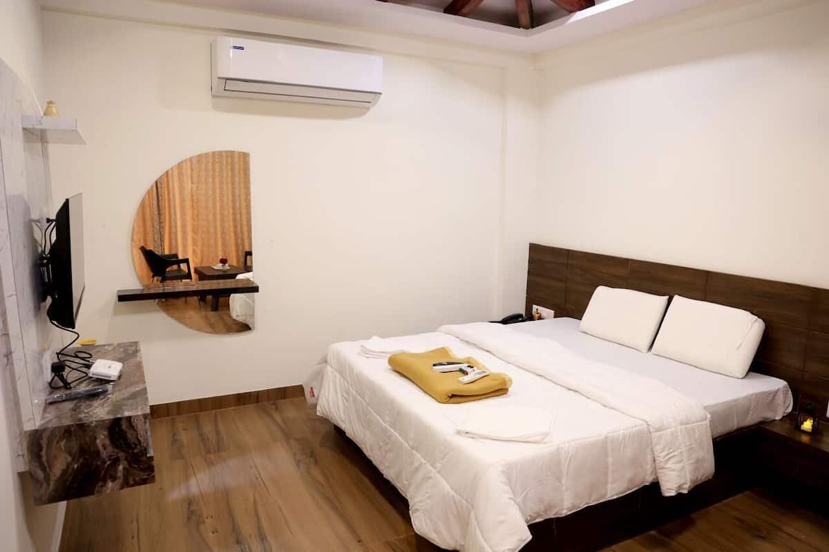 A hotel room with a bed, a TV, an air conditioner, a circular mirror, and a wooden floor.