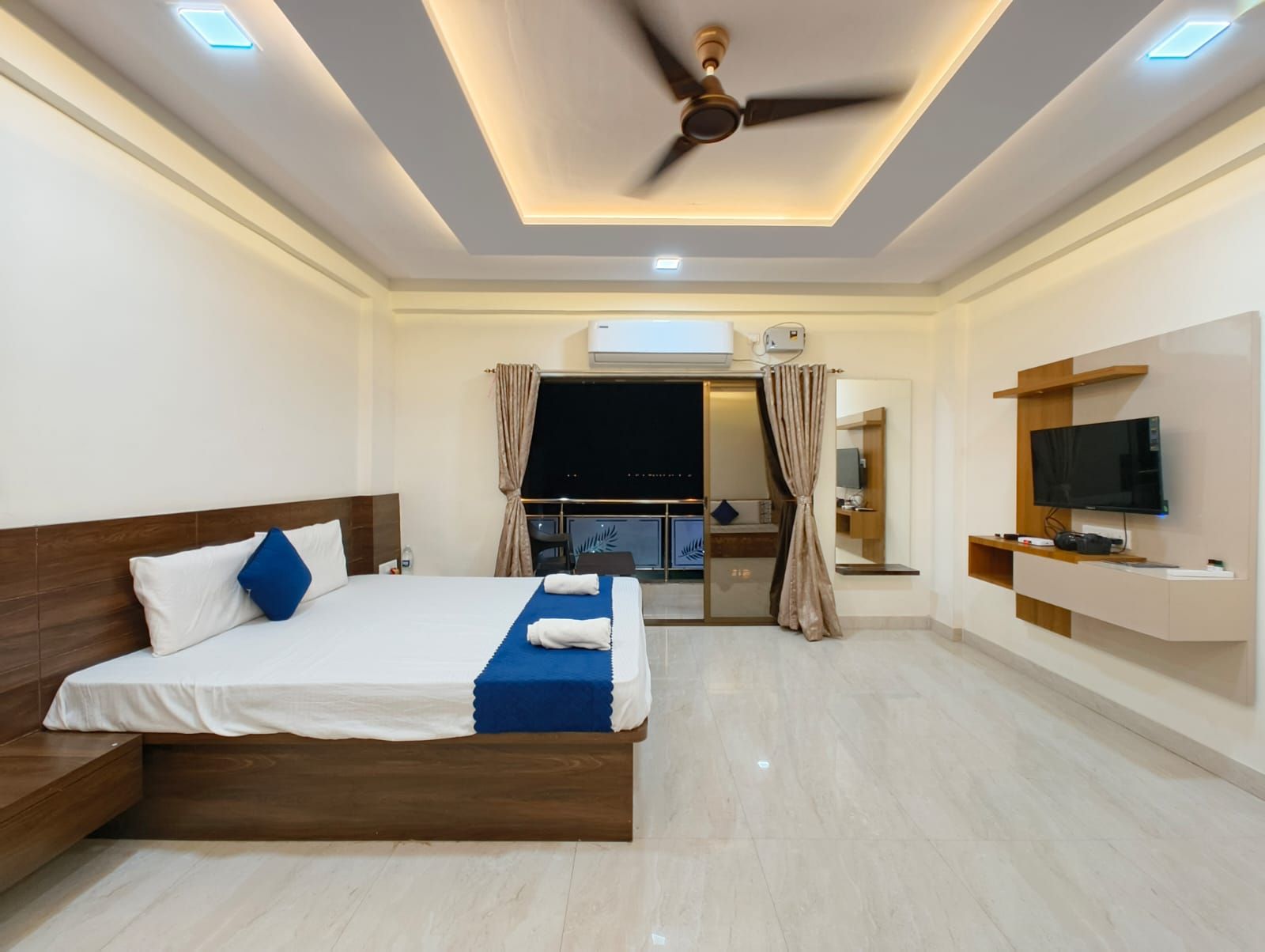 Habitación de hotel con cama, ventilador de techo, televisor y baño visible a través de una puerta de vidrio.
