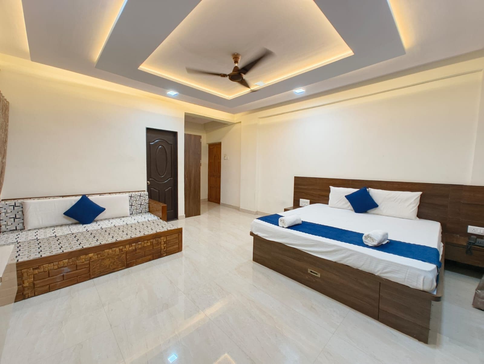 A modern hotel room with a bed, a sofa, a ceiling fan, and a door.