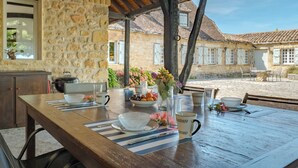 Une table en bois, dressée avec des assiettes, des bols et un vase de fleurs, avec des murs en pierre et un toit en tuiles en arrière-plan.
