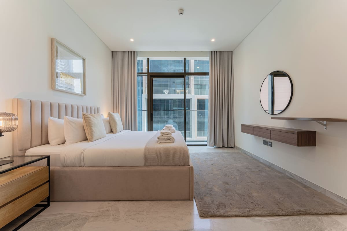 A modern bedroom with a large bed, a bedside table, a mirror, and a view of the city through large windows.