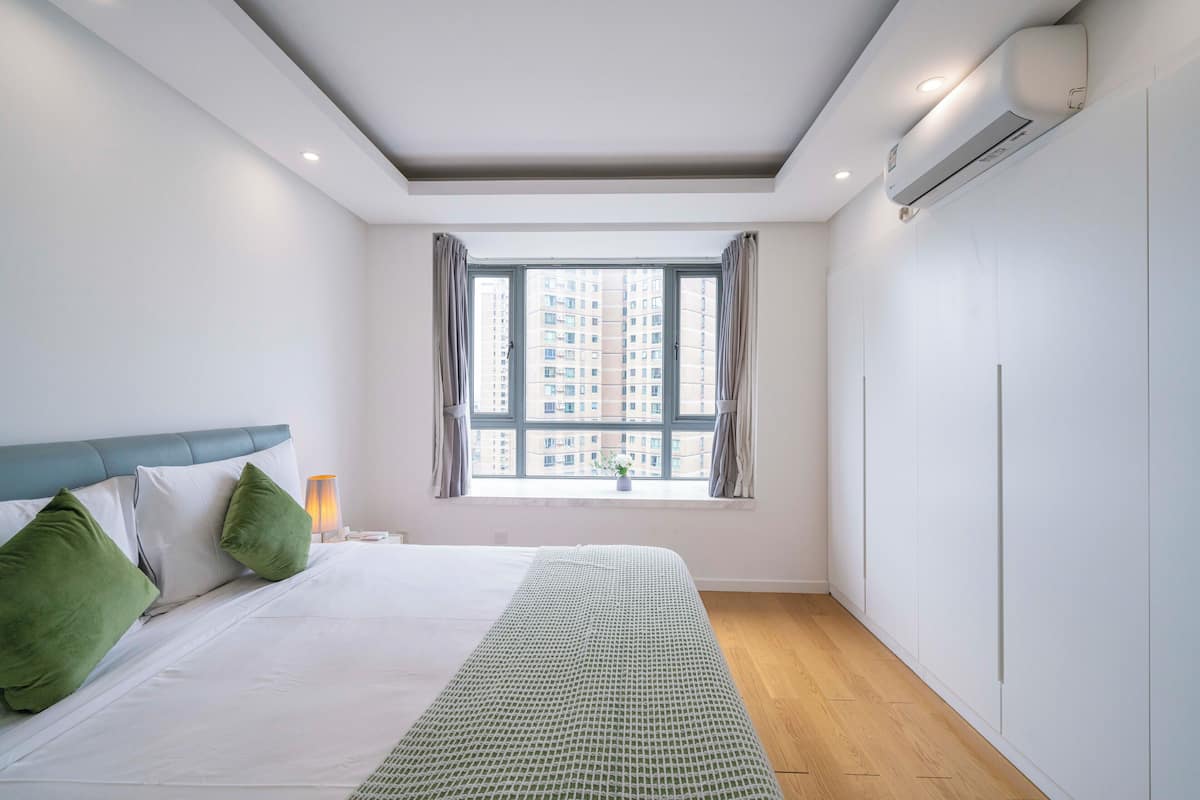 A bedroom with a bed, a window with curtains, a bedside lamp, and an air conditioning unit.