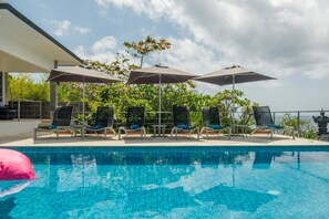 Una piscina con sillas de descanso y sombrillas, rodeada de vegetación y un cielo despejado.