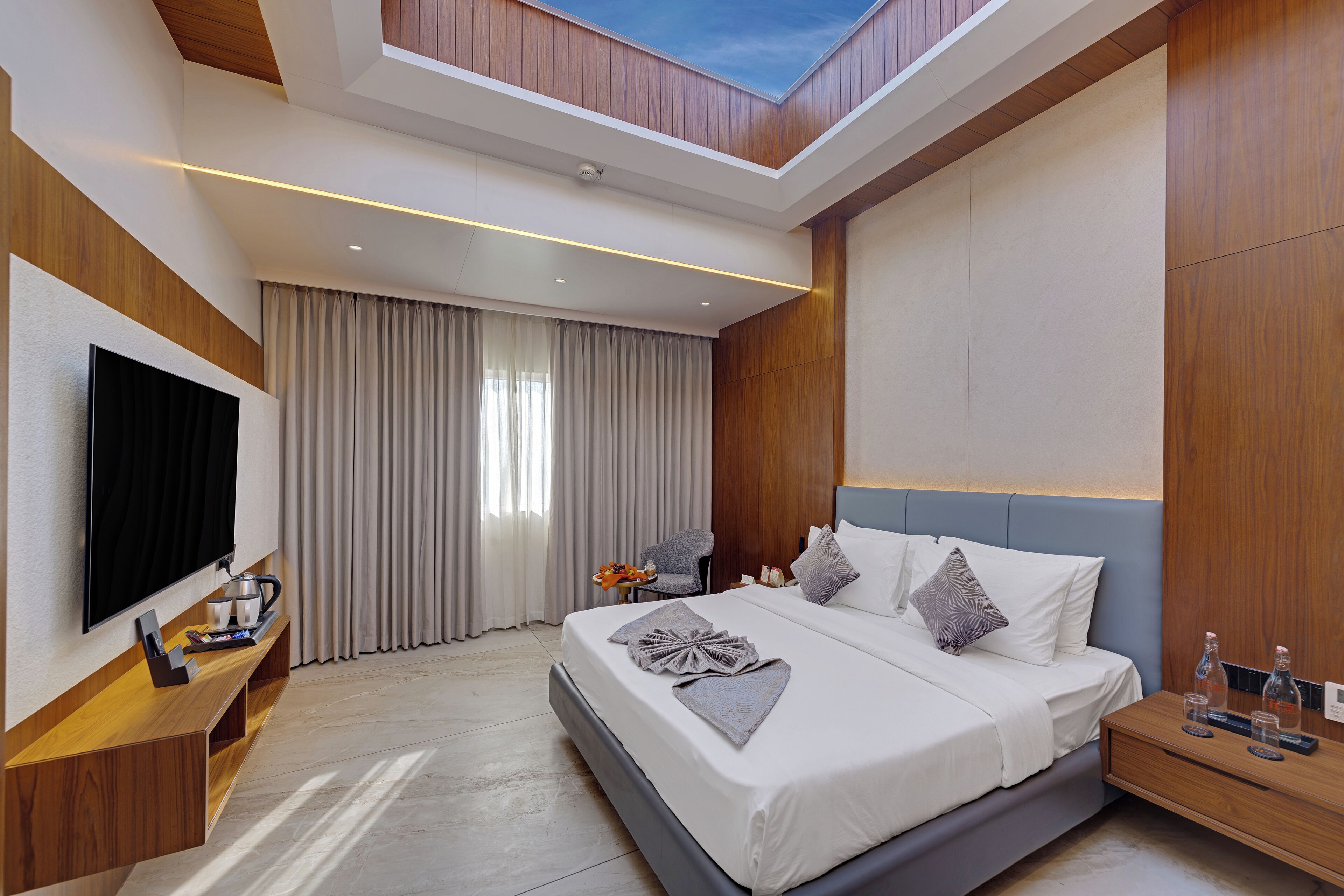 A modern hotel room with a flat-screen TV, a wooden console table, a bed with a grey headboard, and a skylight.