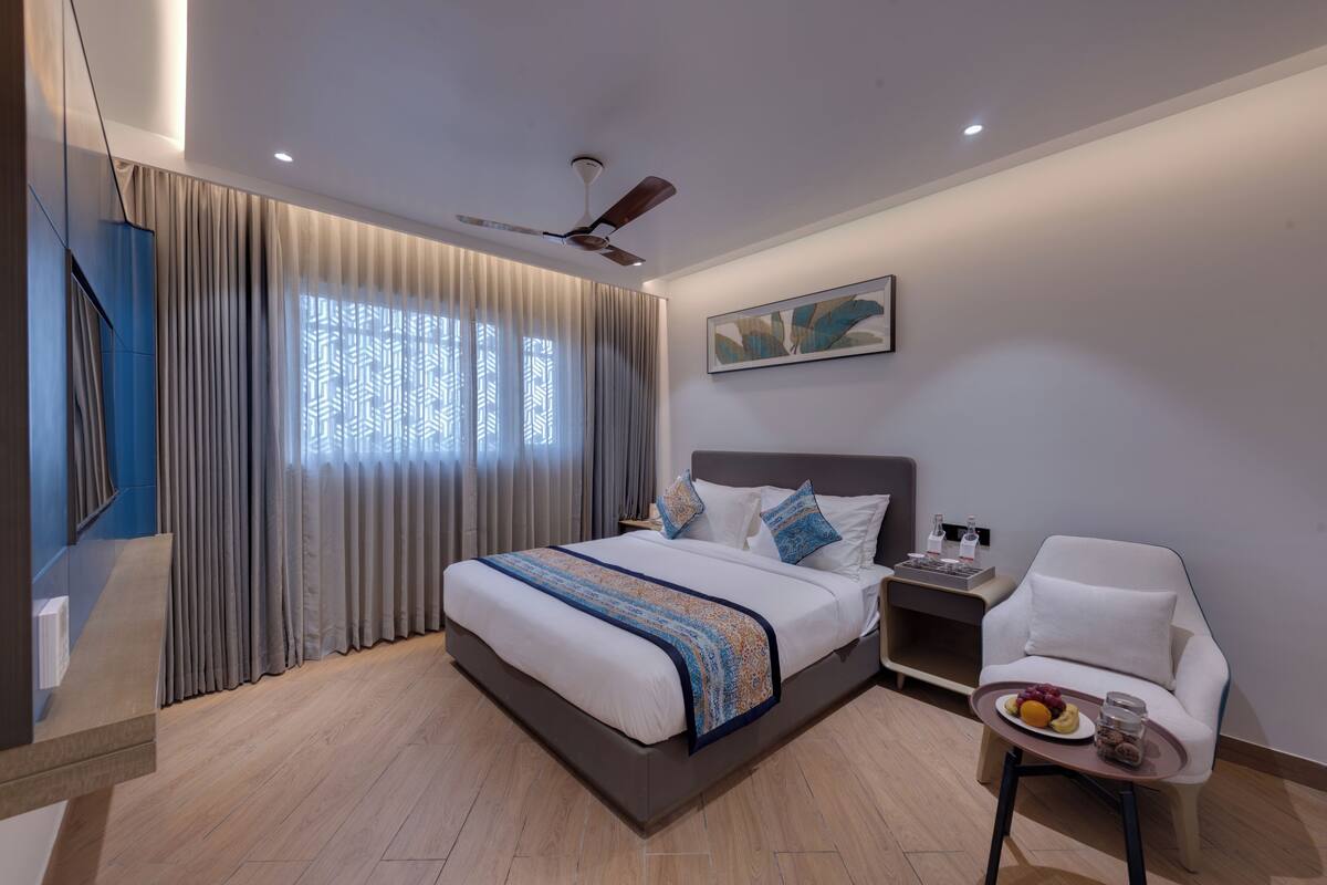 A modern hotel room with a large bed, a bedside table with a lamp, a chair, and a small table with fruit and a glass.