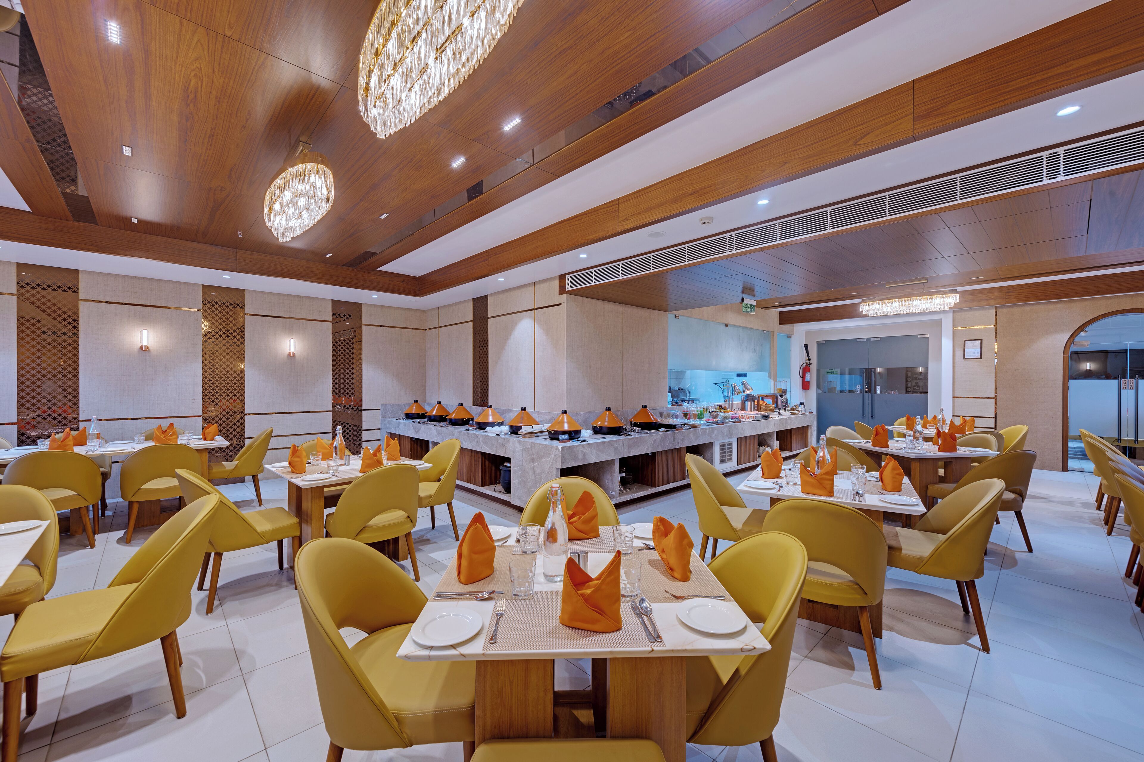 A spacious dining area with yellow chairs and tables, a central island with a kitchen, and a modern chandelier.