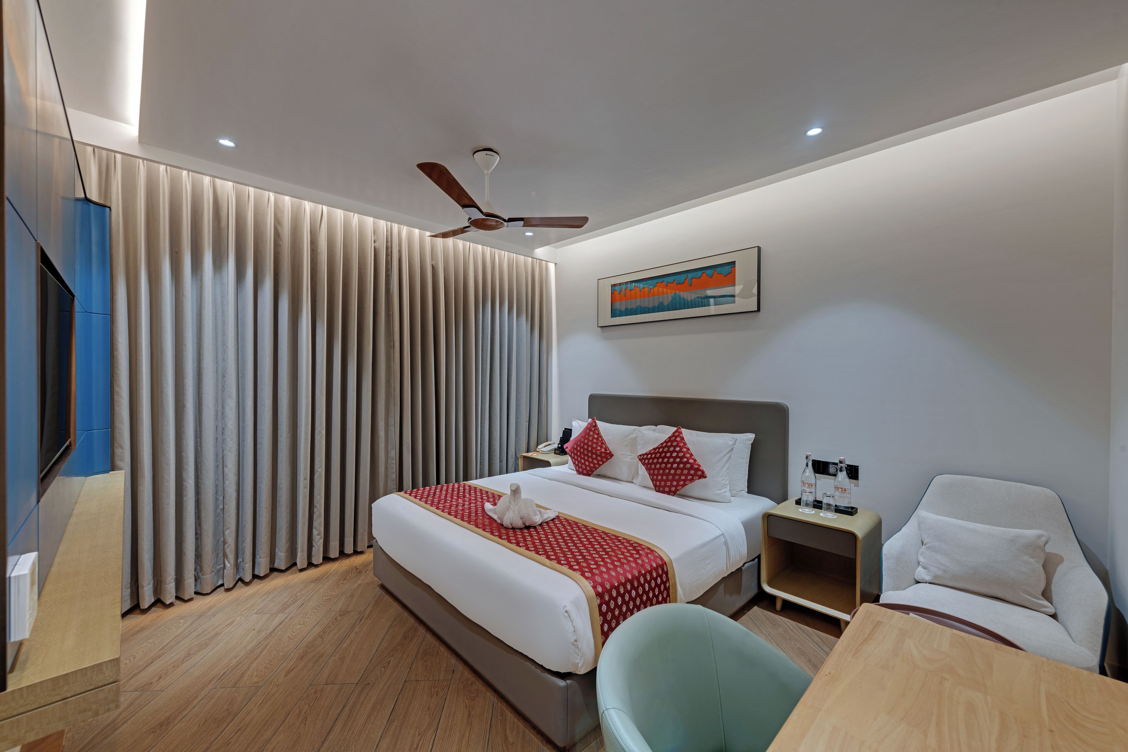 A hotel room with a bed, a desk, a chair, a TV, a ceiling fan, and a painting on the wall.