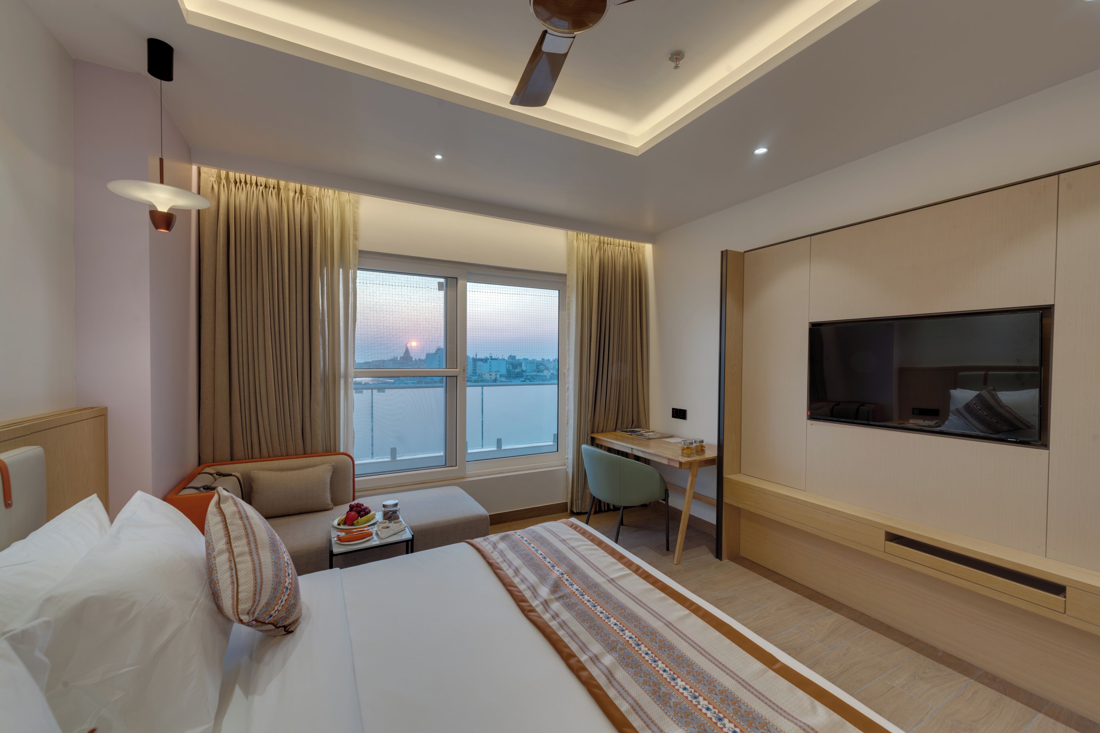 A modern hotel room with a large bed, a desk, a chair, a TV, and a view of the cityscape.