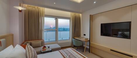 A modern hotel room with a large bed, a desk, a chair, a TV, and a view of the cityscape.