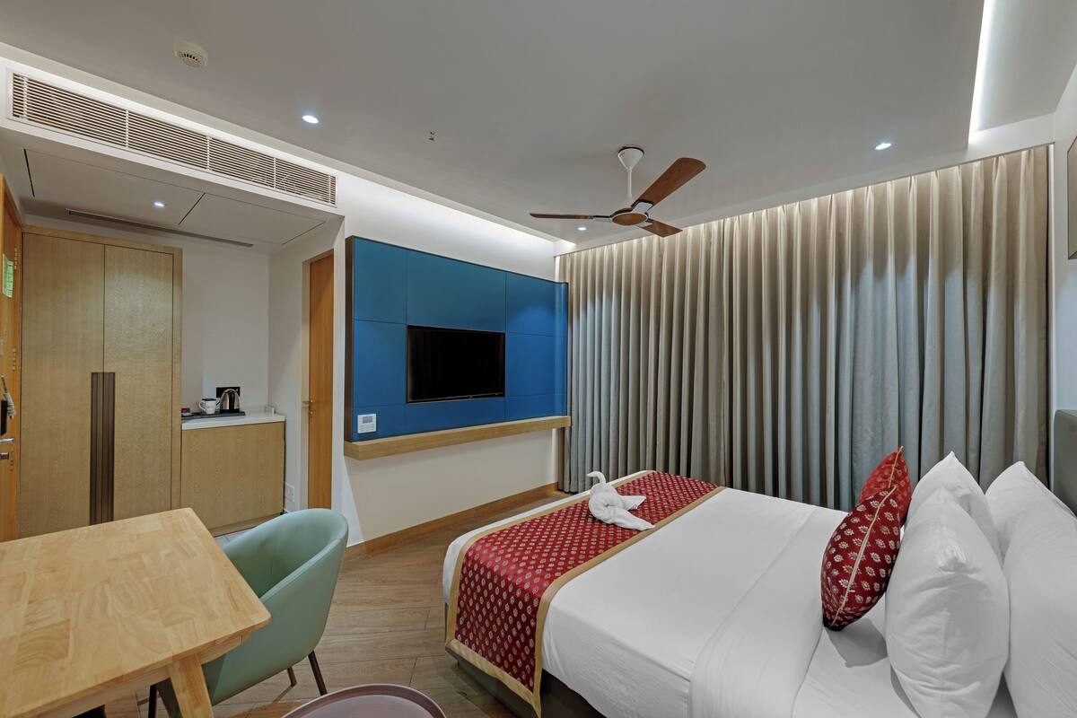A hotel room with a bed, a TV mounted on the wall, a ceiling fan, and a wooden table.
