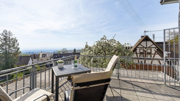 A balcony with a table and chairs, a view of buildings, and a tree with white flowers.
