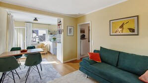 A living room with a green sofa, a dining table with chairs, a kitchen area, and a framed picture on the wall.