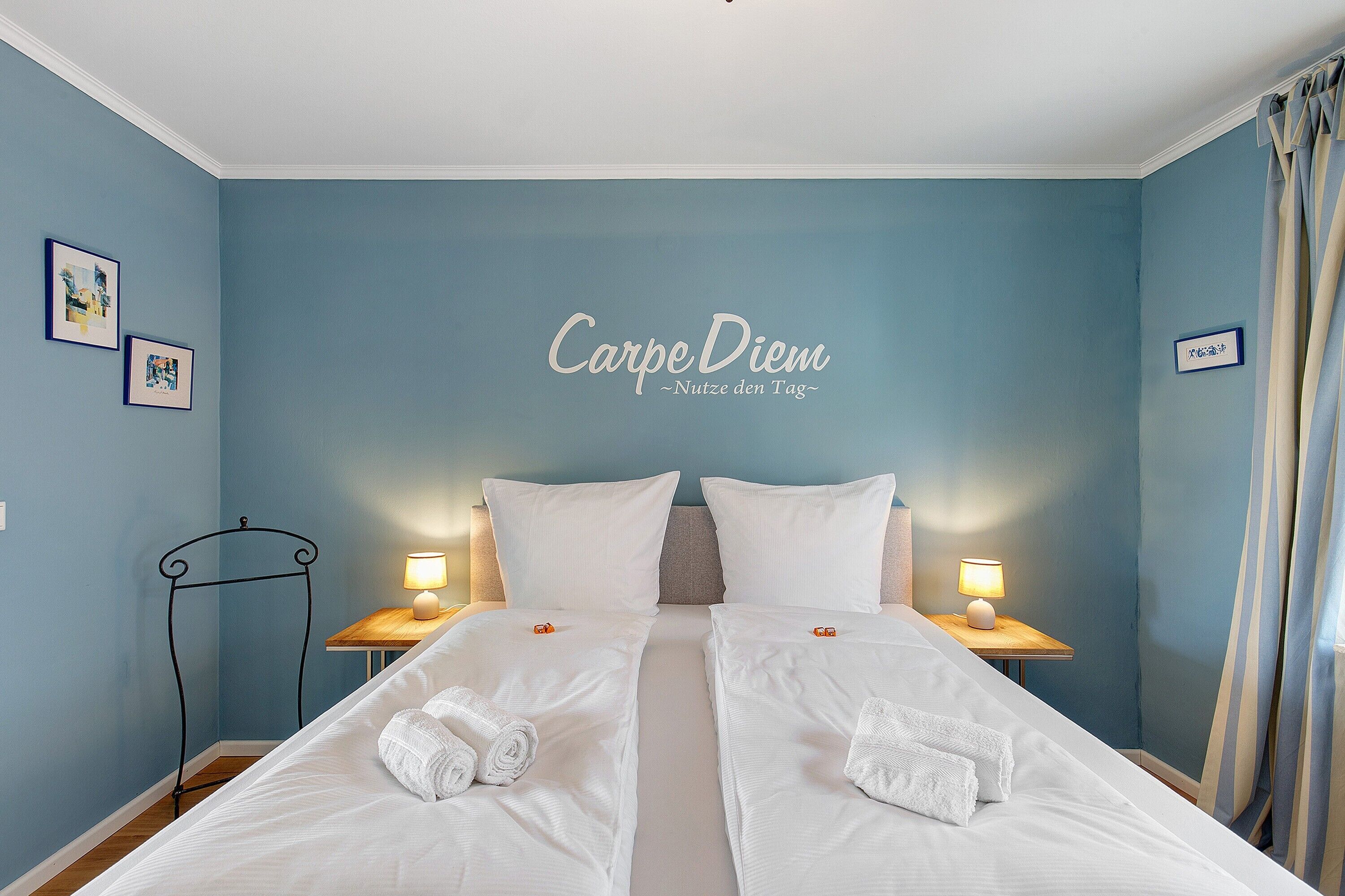 Ein Doppelbett mit weißen Leinen und Handtüchern, zwei Nachttische mit Lampen, ein Metallgestell und eine Wand mit Text.