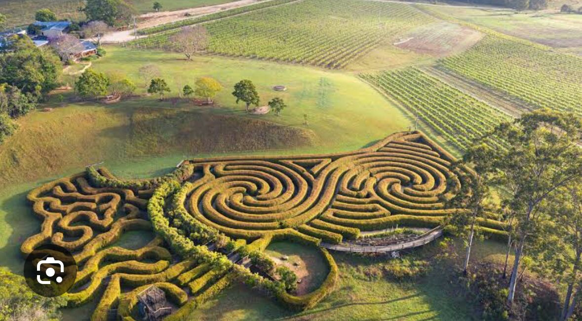 Ein weitläufiger, verschlungener Labyrinthgarten mit zahlreichen Pfaden und einer zentralen, kreisförmigen Fläche, umgeben von offenen Feldern und vereinzelten Bäumen.