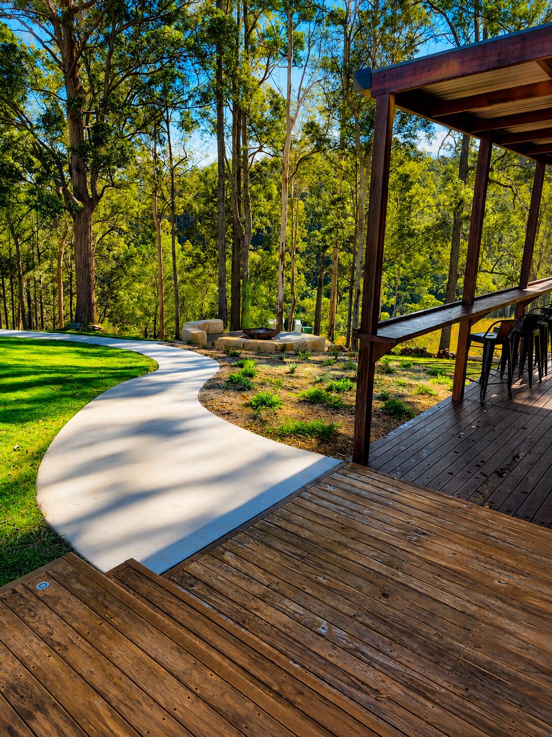 Una terraza de madera con un sendero curvo que lleva a una zona de estar bajo una pérgola.