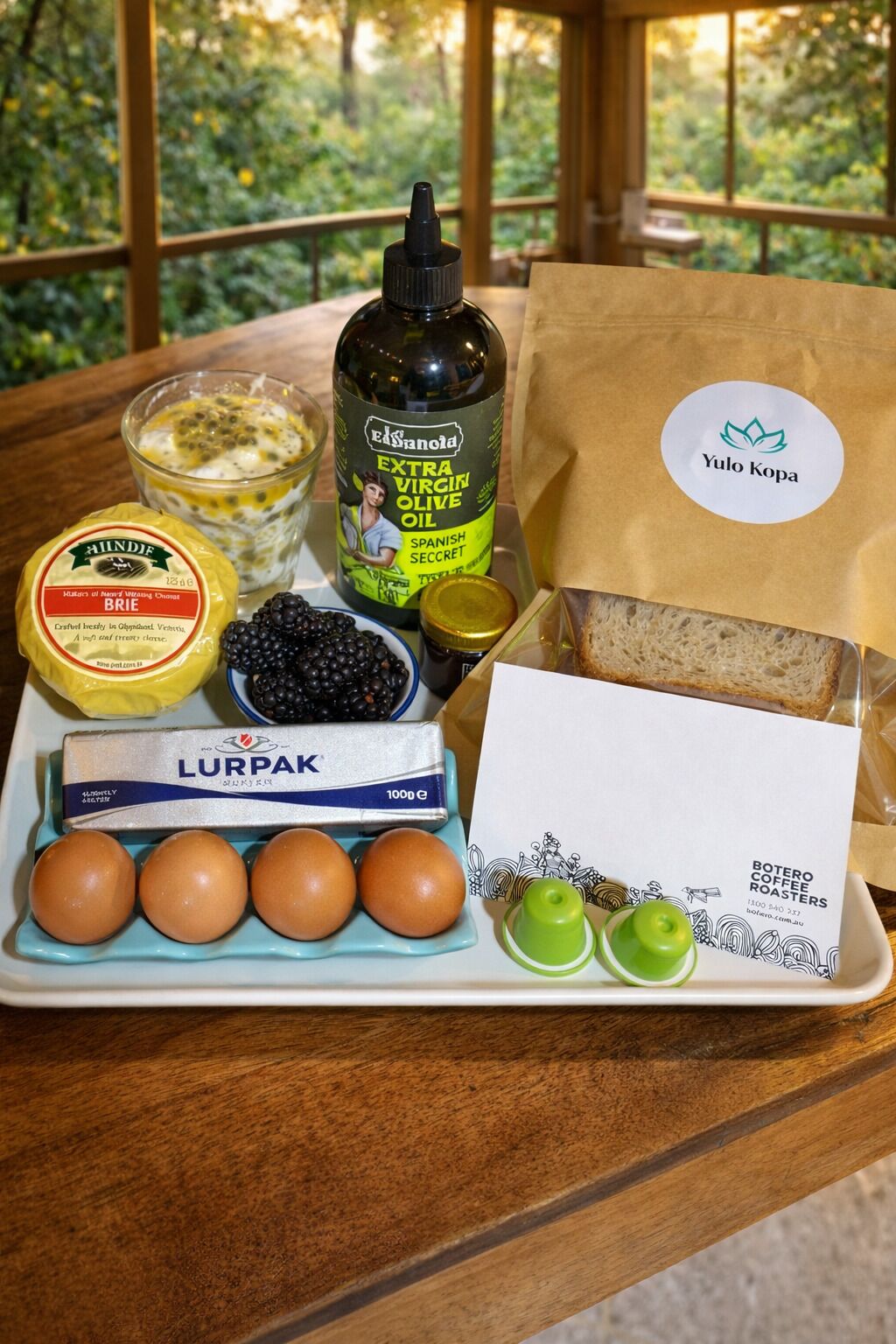 A table with a bottle of extra virgin olive oil, a jar of blackberries, a pack of Lurpak butter, a carton of eggs, and a package of Botero coffee roasters.