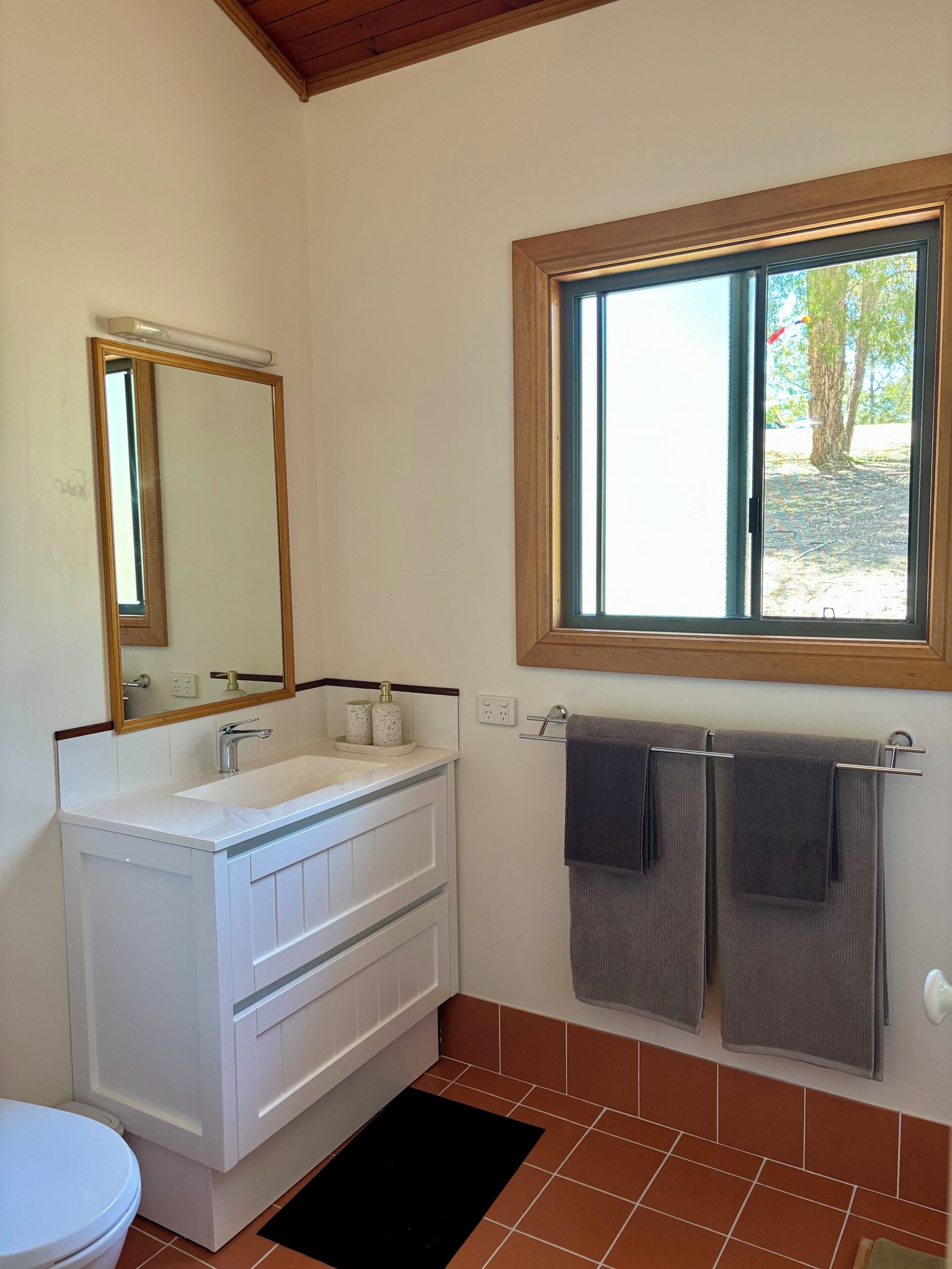 Un baño con un lavamanos blanco, un espejo, una ventana y dos toallas colgadas en un perchero.