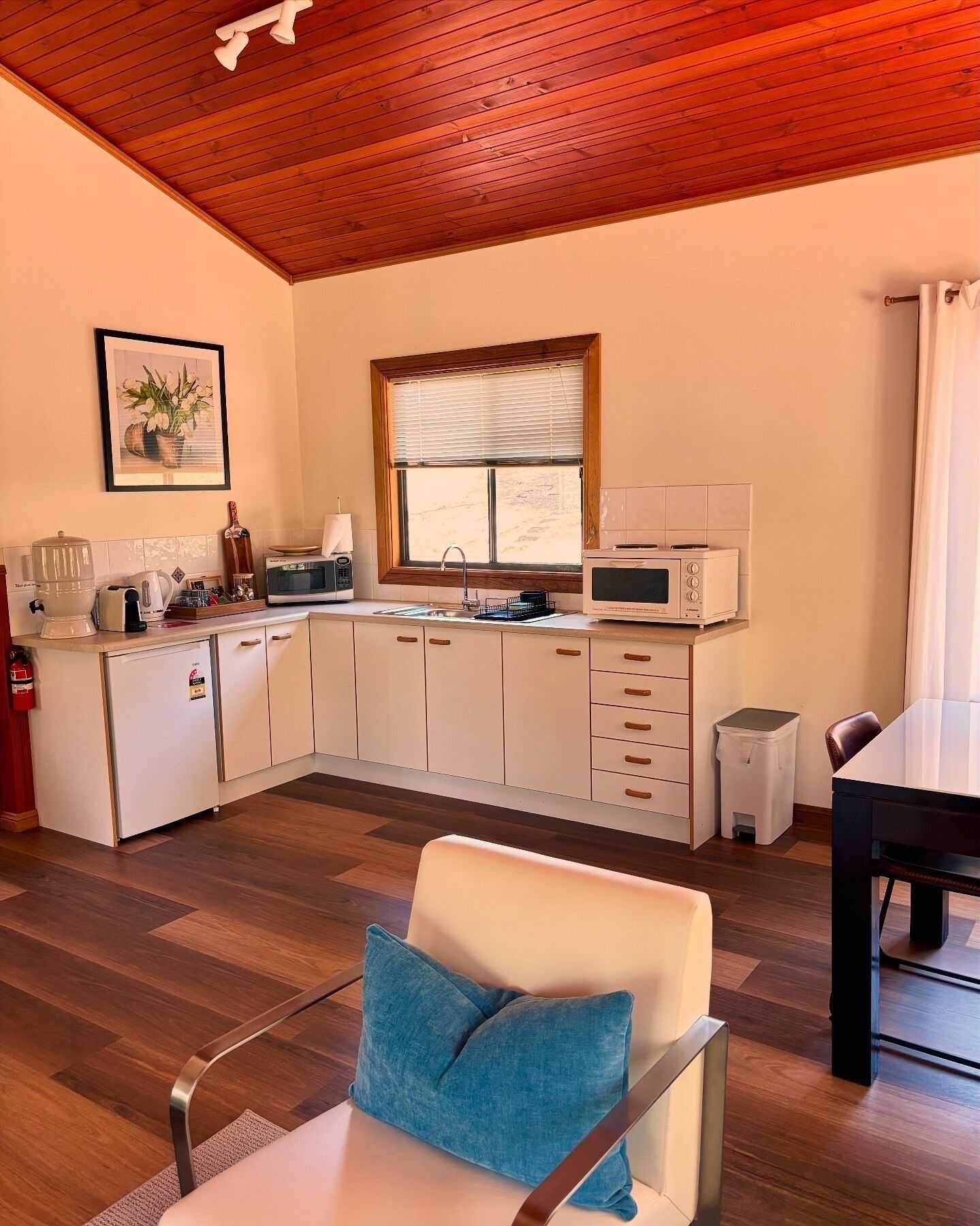 A compact kitchen with white cabinets, a wooden ceiling, a window with blinds, a microwave, and a sink.