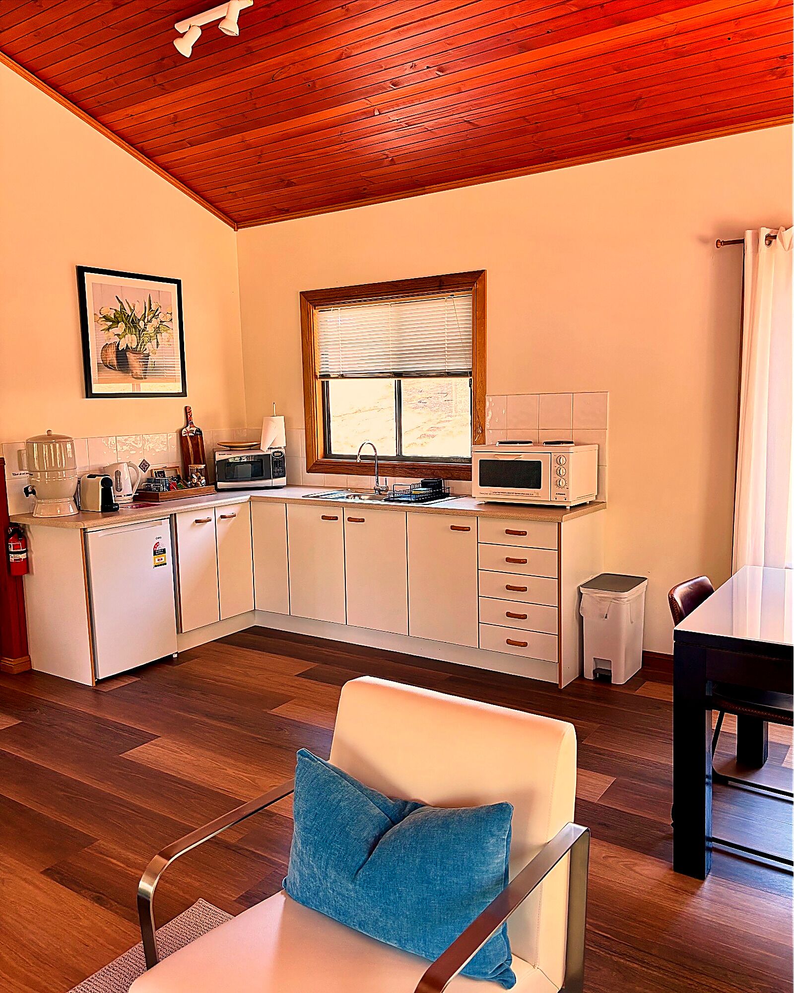 A compact kitchen with white cabinets, a microwave, and a sink. There is a framed picture on the wall and a window with blinds.