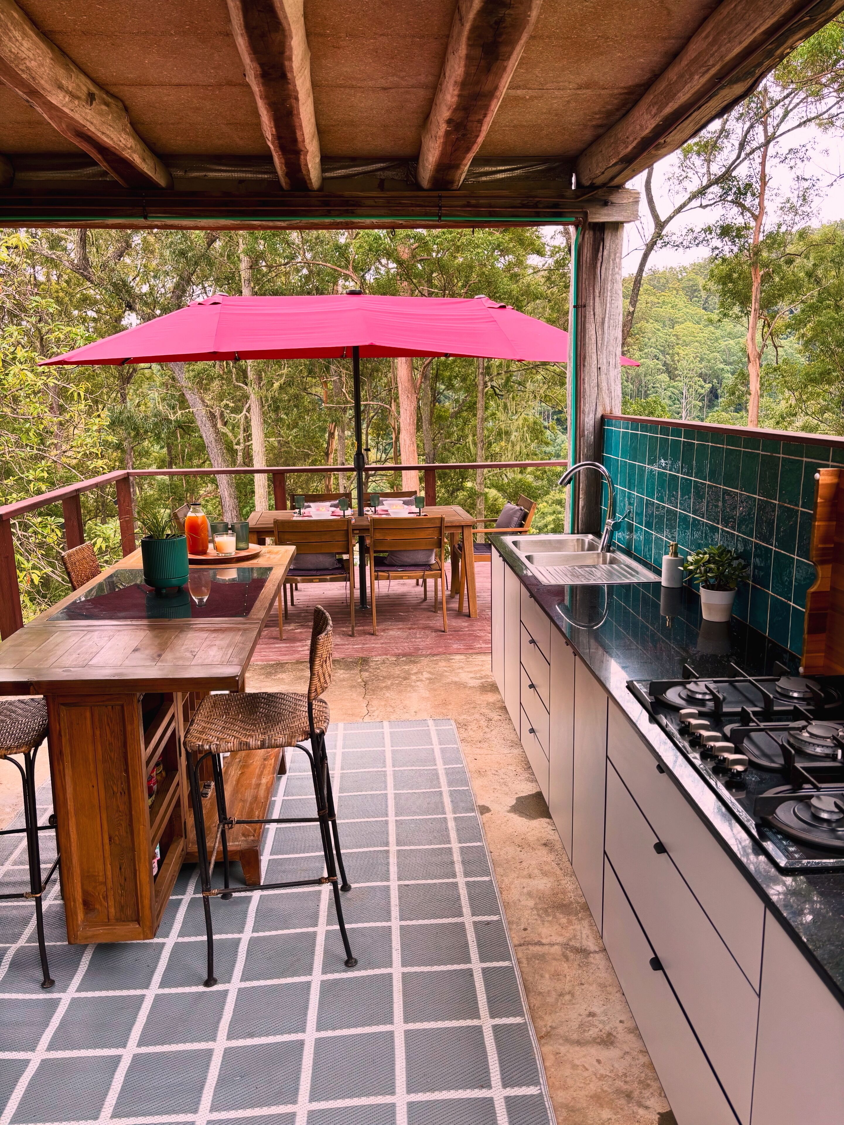 Una cocina al aire libre techada con mesa y sillas, una sombrilla roja y vistas del bosque circundante.