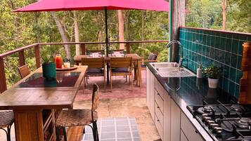 Una cocina al aire libre techada con mesa y sillas, una sombrilla roja y vistas del bosque circundante.