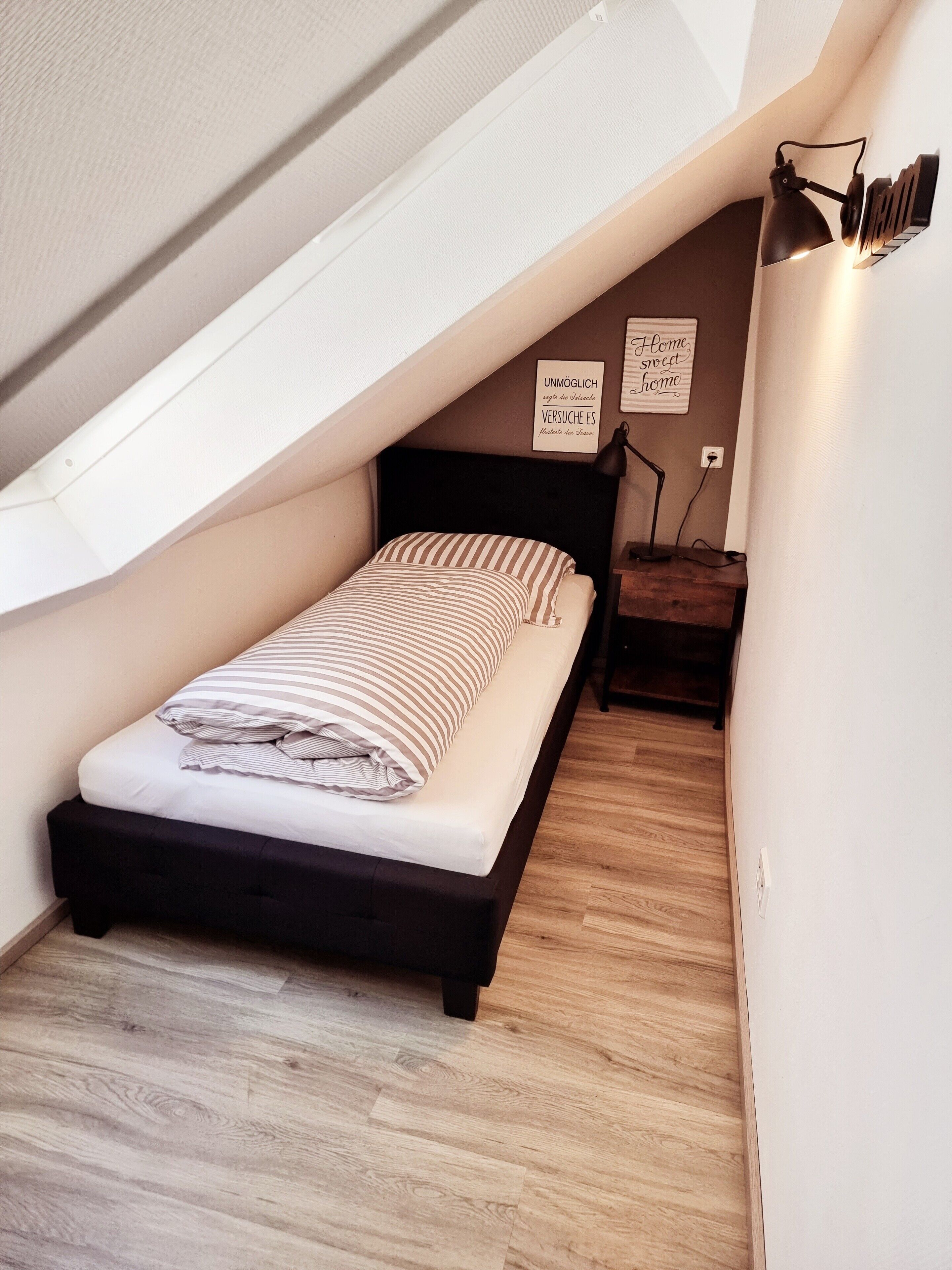 Une petite chambre à coucher accueillante, avec un plafond en pente, un lit avec une literie à rayures, une table de chevet et une lampe fixée au mur.