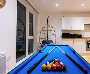 Une table de billard avec des billes, un fauteuil en osier, un coin cuisine et une fenêtre donnant sur un beau paysage.