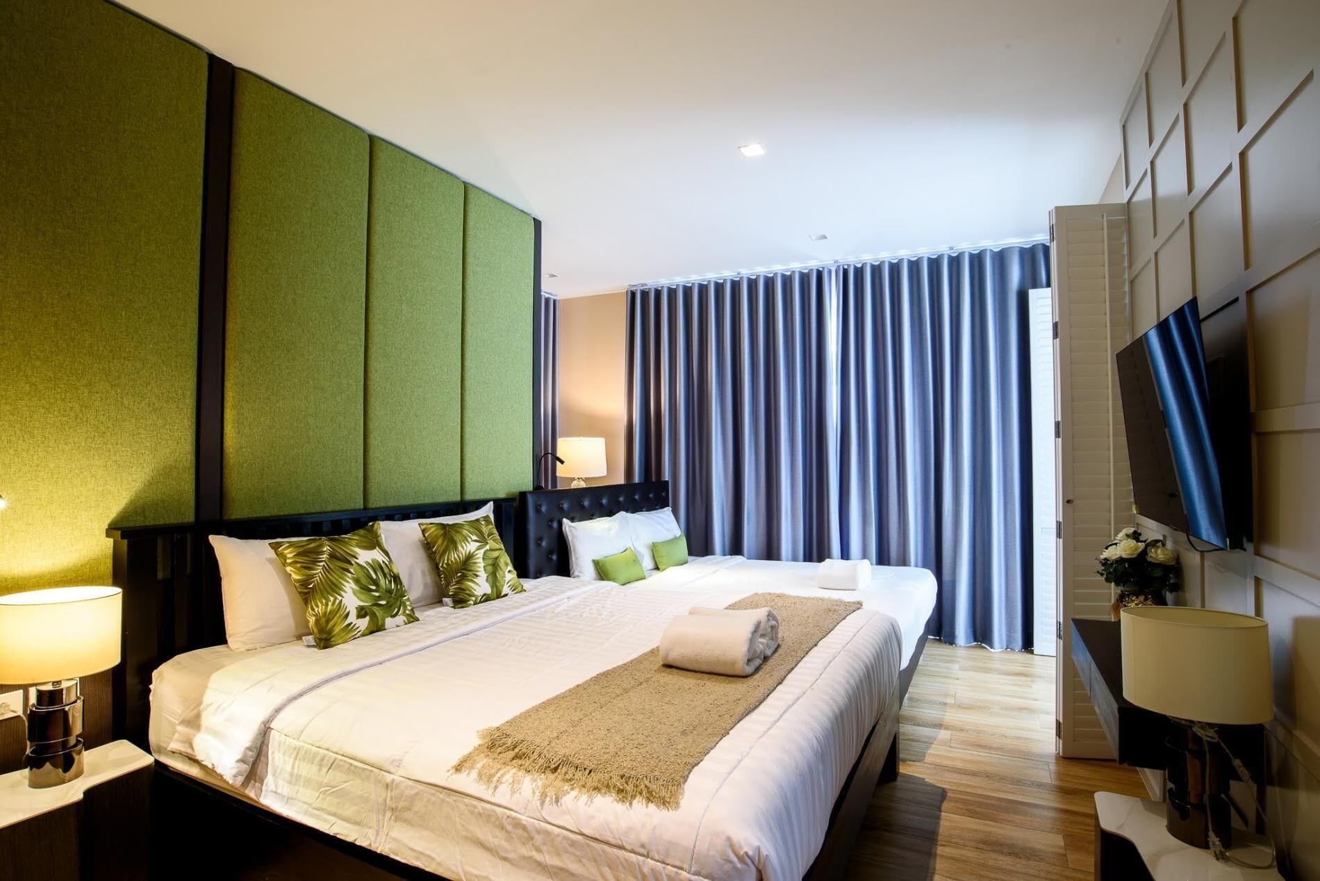 A hotel room with a large bed, green paneling on the wall, a flat-screen TV, and a large window with curtains.