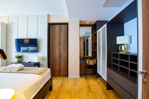A hotel room with a bed, a TV mounted on the wall, a wooden door, a desk with a chair, and a wardrobe with shelves. - Pattaya Inara Exclusive Pool Villa PI5 (Pattaya)