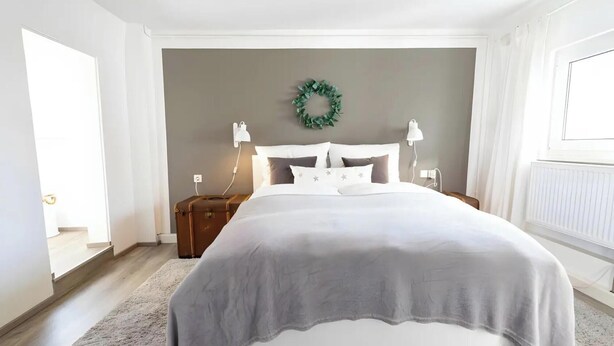 A bedroom with a bed, nightstands, a wall-mounted lamp, a power outlet, a suitcase, and a wreath. - Apartment 'Snug' with Wi-Fi (Neuenstein)